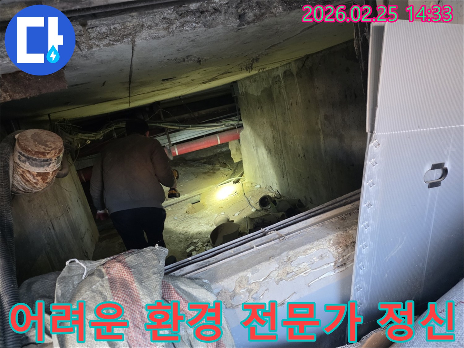 누수 전문 엔지니어가-어둡고 좁은 어려운 환경 속에서-손전등을 비춰가며-누수 지점을 찾아내기 위해-최선을 다하는 전문가 정신을-보여주는 모습입니다.