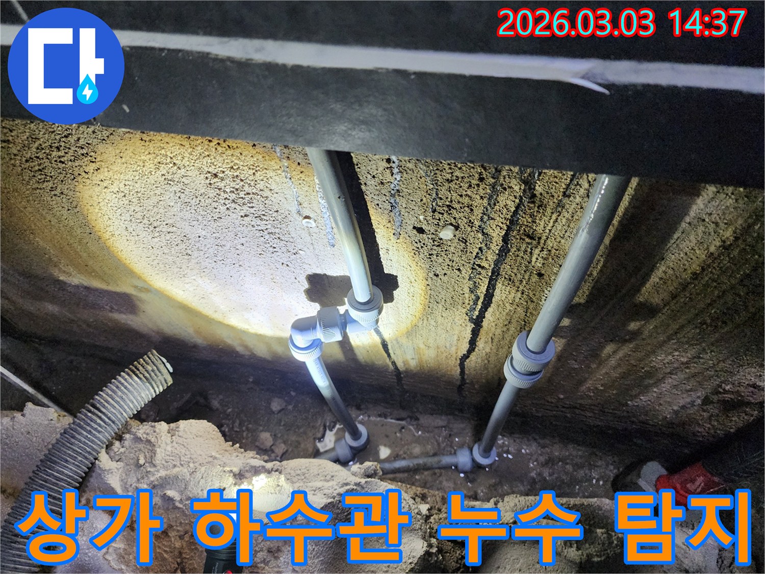 어두운 상가 지하에서-손전등으로 비춘-하수 배관의-연결부 누수를-전문가가 확인하는 장면입니다.