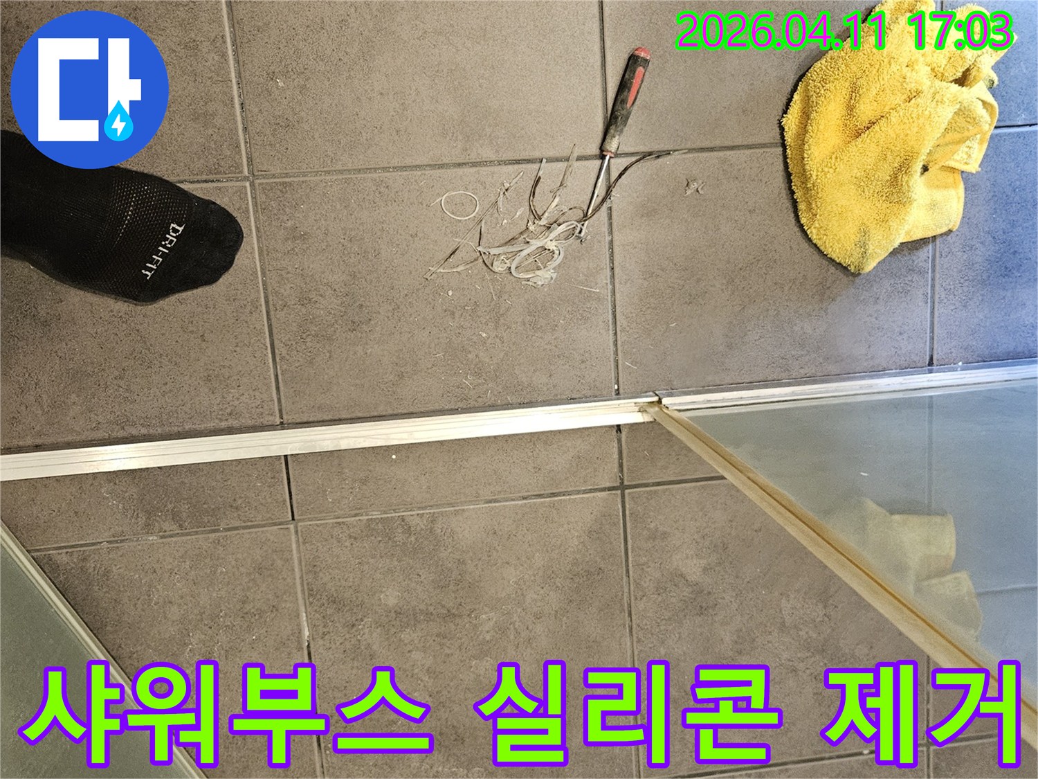 욕실 바닥 타일 위-노란 수건과 공구-그리고 샤워부스 주변의 낡은 실리콘을 제거하는 작업 모습