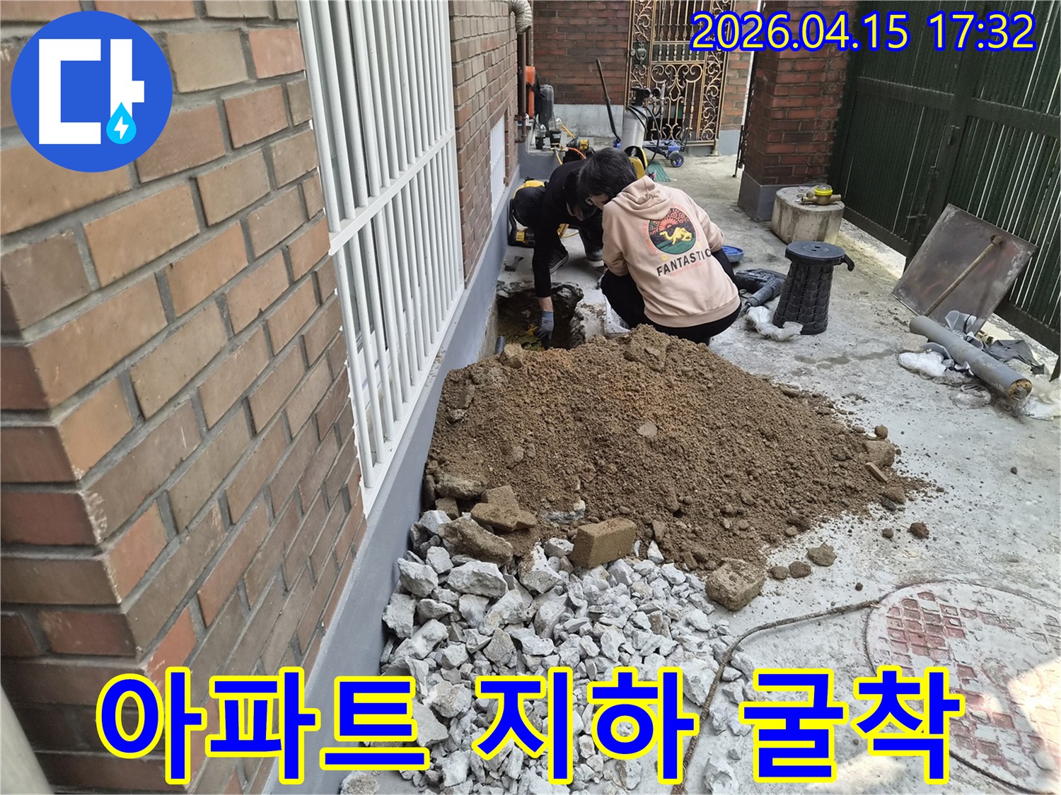 두 명의 작업자가-아파트 건물 옆에서-지하 누수 지점을 찾아-땅을 파는 현장