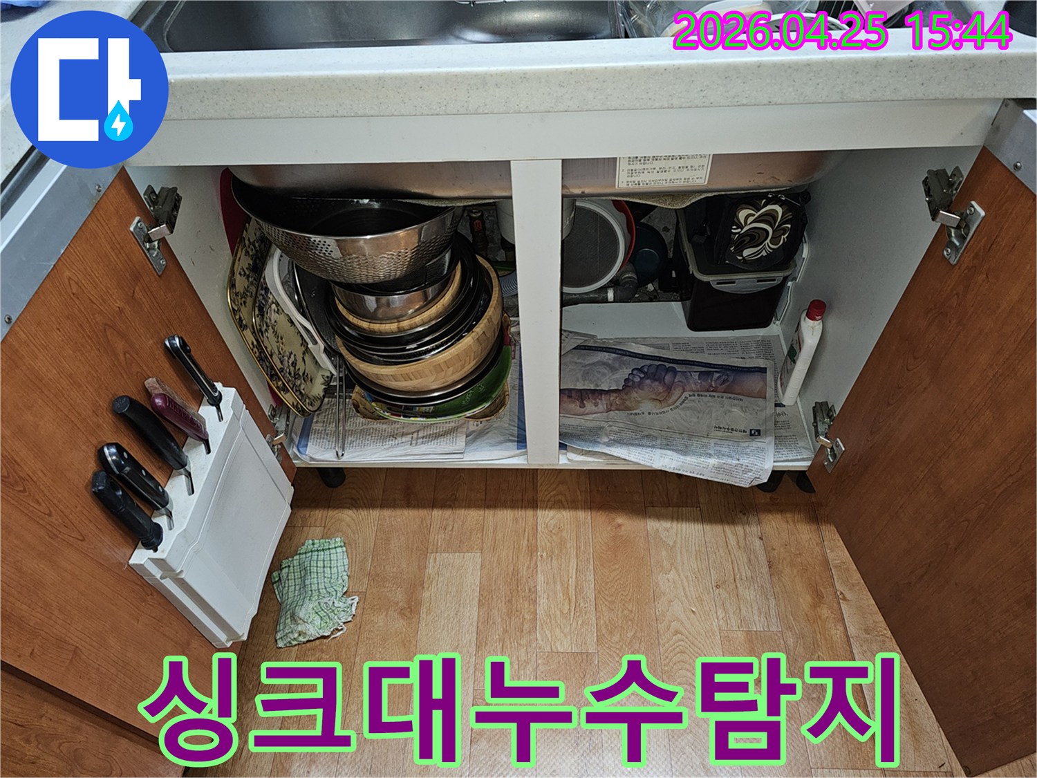 싱크대 하부장 내부-누수 점검을 위해-그릇과 배관이 보이고 바닥에 신문지가 깔려 있습니다.