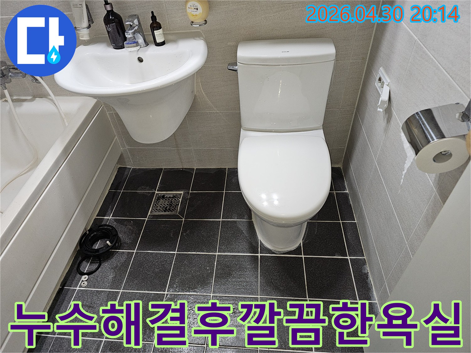 누수 수리 작업이 완료된 후의 아파트 욕실을 보여주는 이미지입니다-하얀색 변기와 세면대가 깨끗하게 설치되어 있으며-바닥의 검은색 타일이 정돈된 모습을 자랑합니다