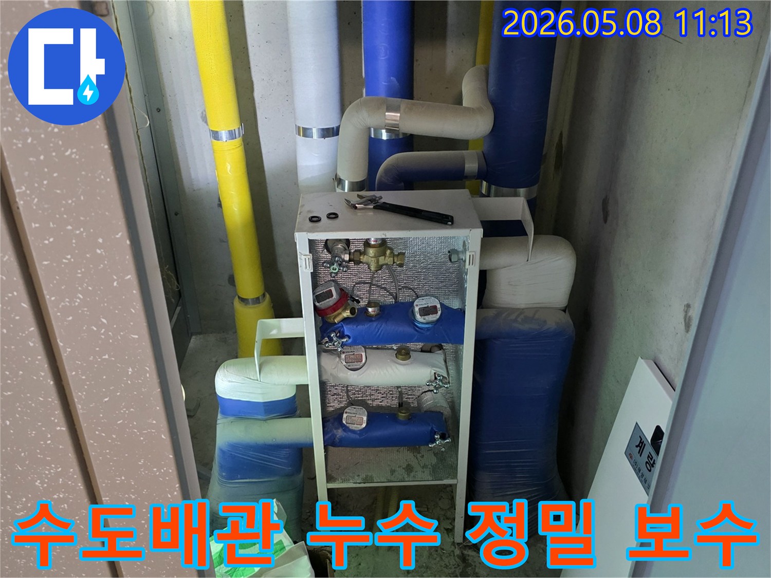 이 이미지는 건물의 수도 공급 시스템이 모여 있는 공간으로-다양한 색상의 단열재로 감싸진 배관과-여러 개의 수도 계량기-그리고 작업 도구가 배치되어 있습니다-이는 수도 누수 보수의 중요성을 나타냅니다