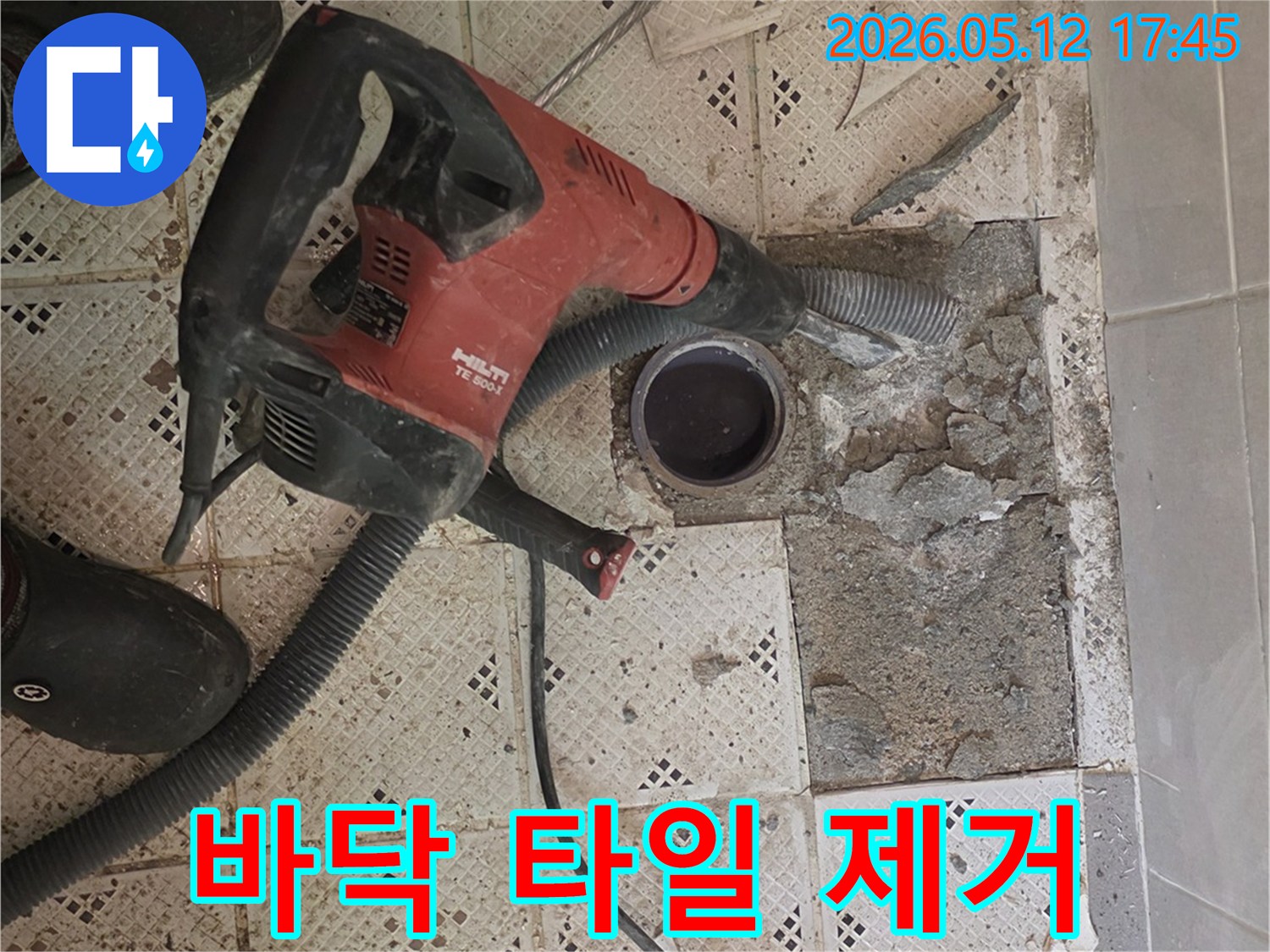 누수 공사를 시작하기 위해-바닥 타일을 제거하는-강력한 해머 드릴과-작업자의 발이 보이는-현장 사진입니다.