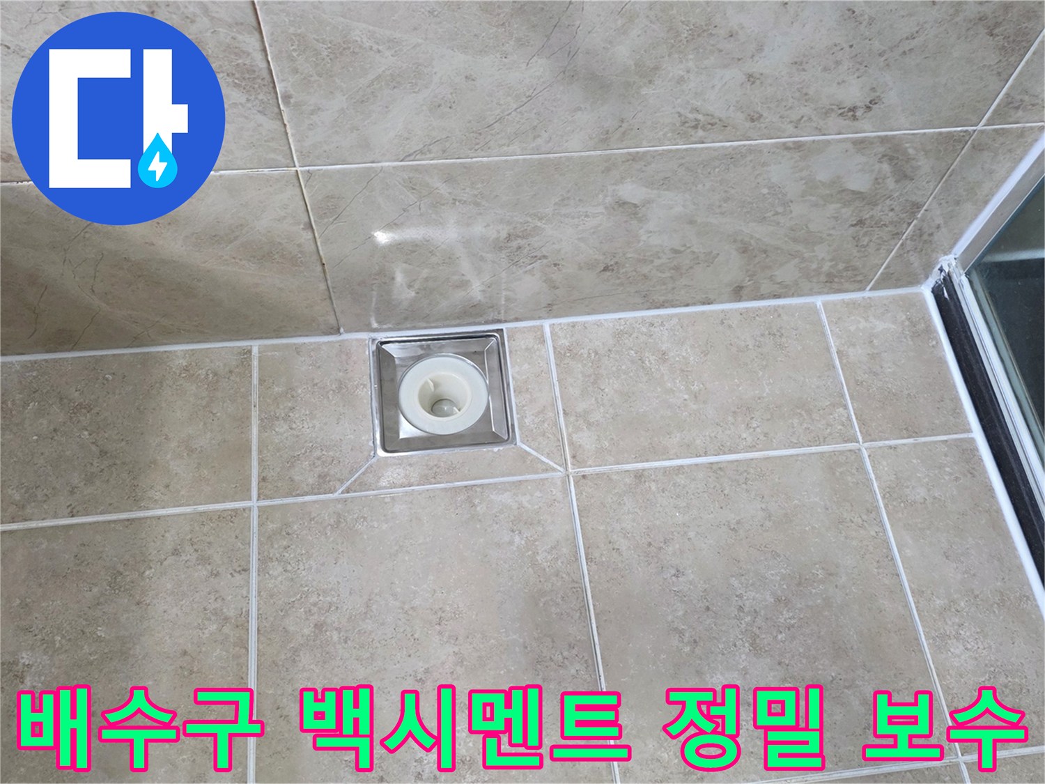 베이지색 타일 바닥에 설치된-사각형 금속 배수구의 모습으로-주변 백시멘트의 마감 상태를 보여줍니다