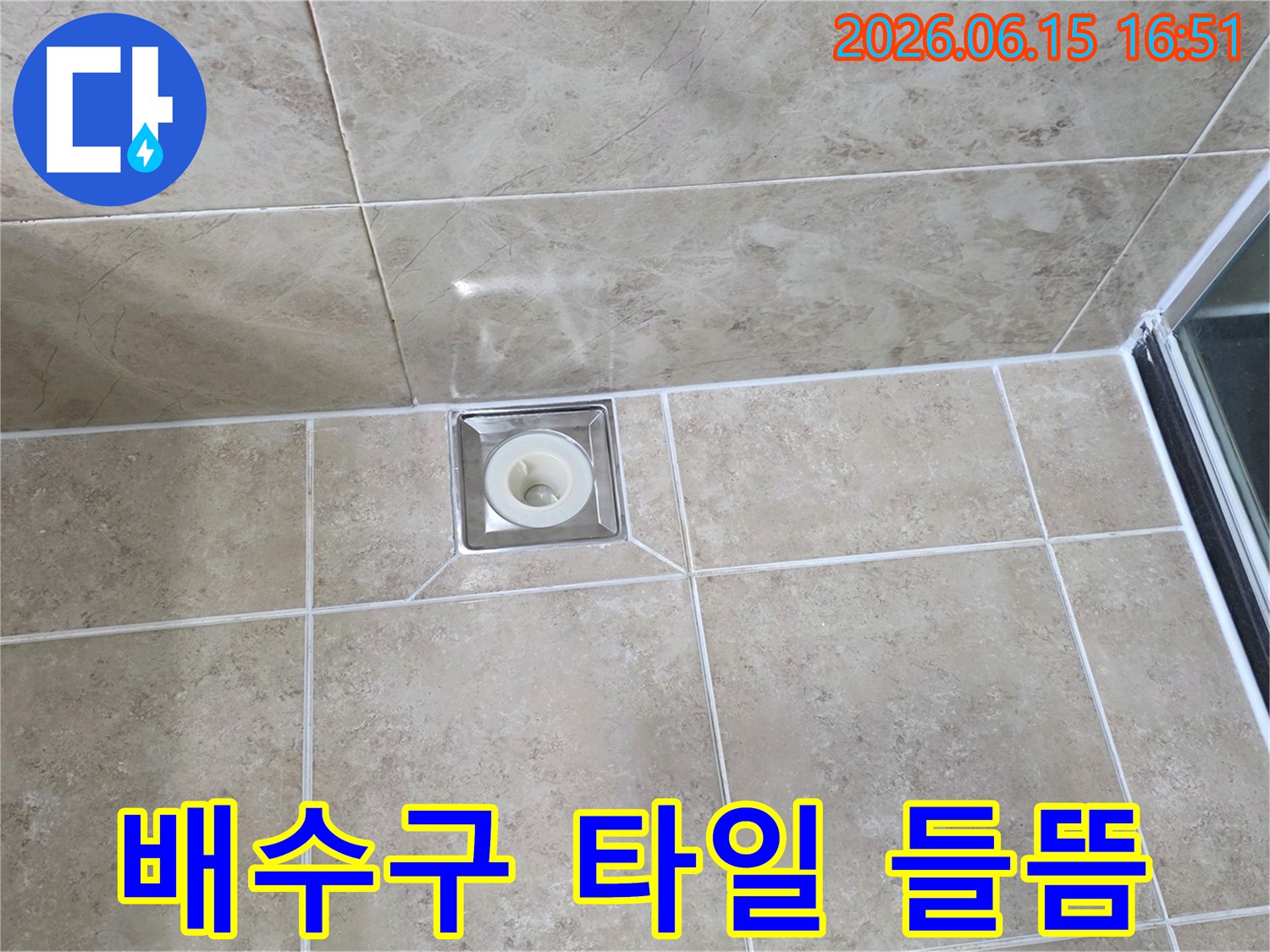베이지색 타일 바닥에 위치한-사각형 금속 배수구의 모습으로-주변 타일의 접합부와-줄눈의 상태를 상세히 보여줍니다