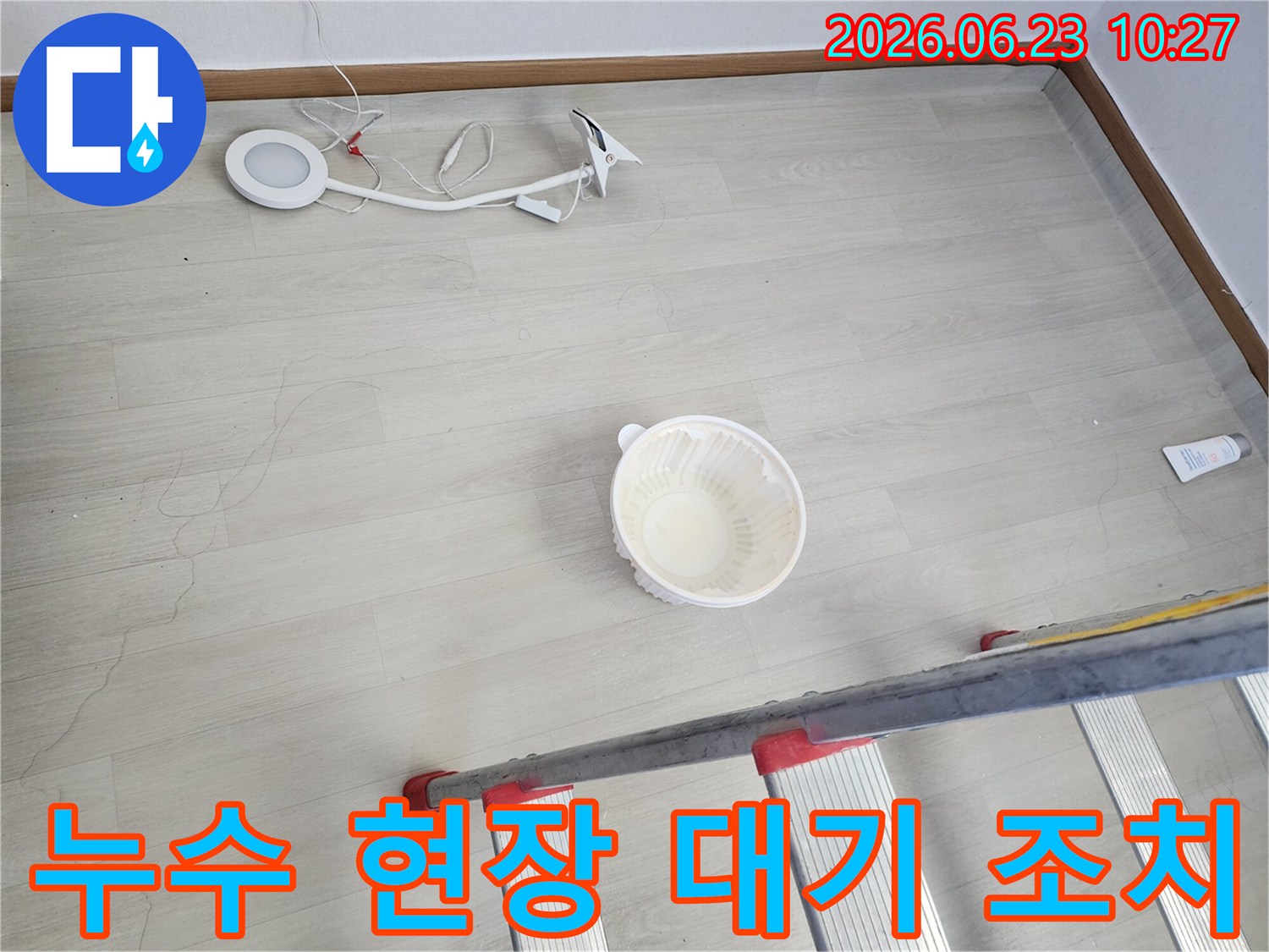 천장에서 물이 떨어져 바닥에 놓인 물받이 그릇과-누수 지점 확인 및 작업을 위한 사다리-조명 장비가 함께 보이는-누수 현장 준비 단계의 모습입니다