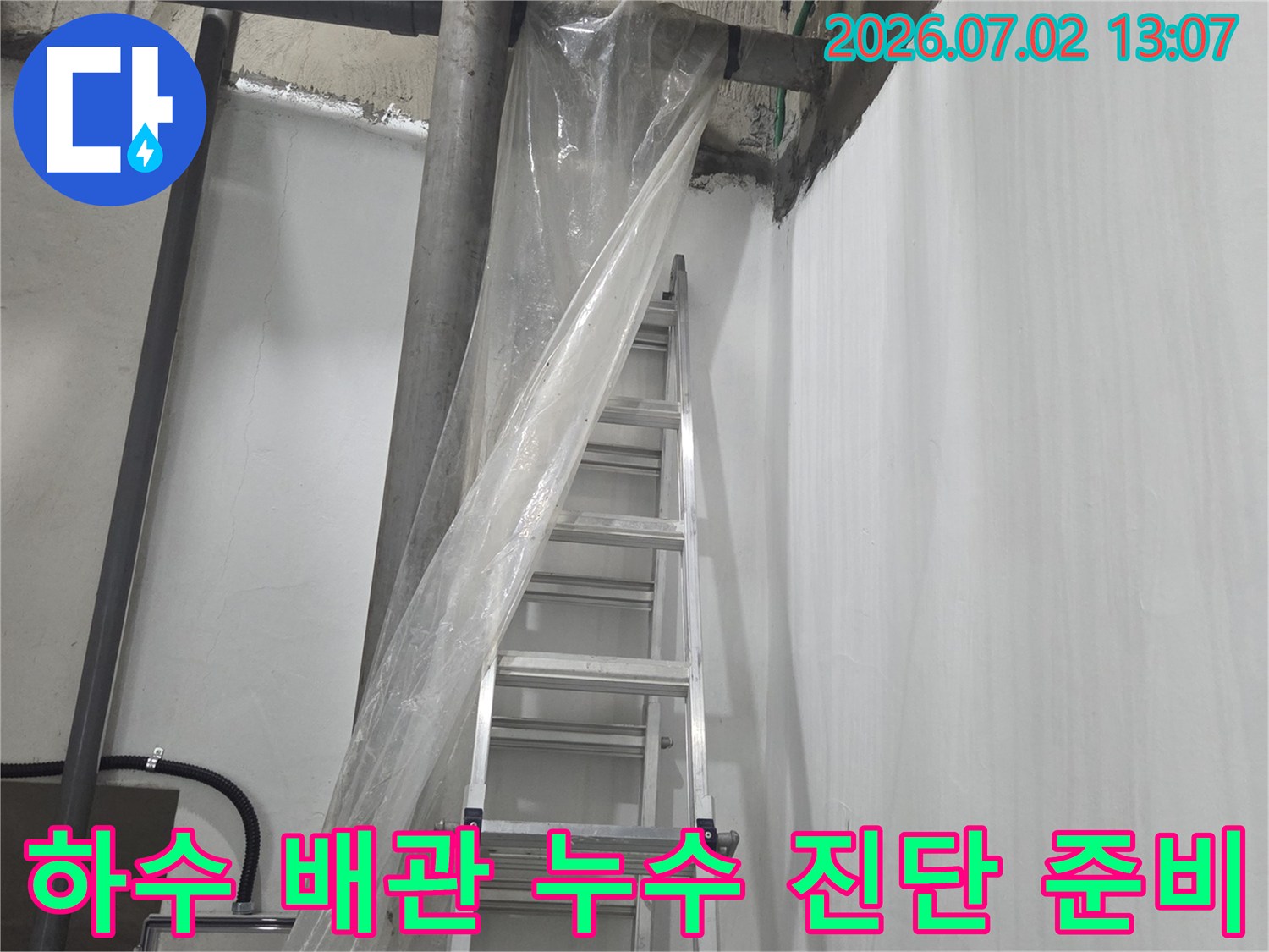 지하 천장의 회색 하수 배관들과 그 아래 설치된 작업용 알루미늄 사다리