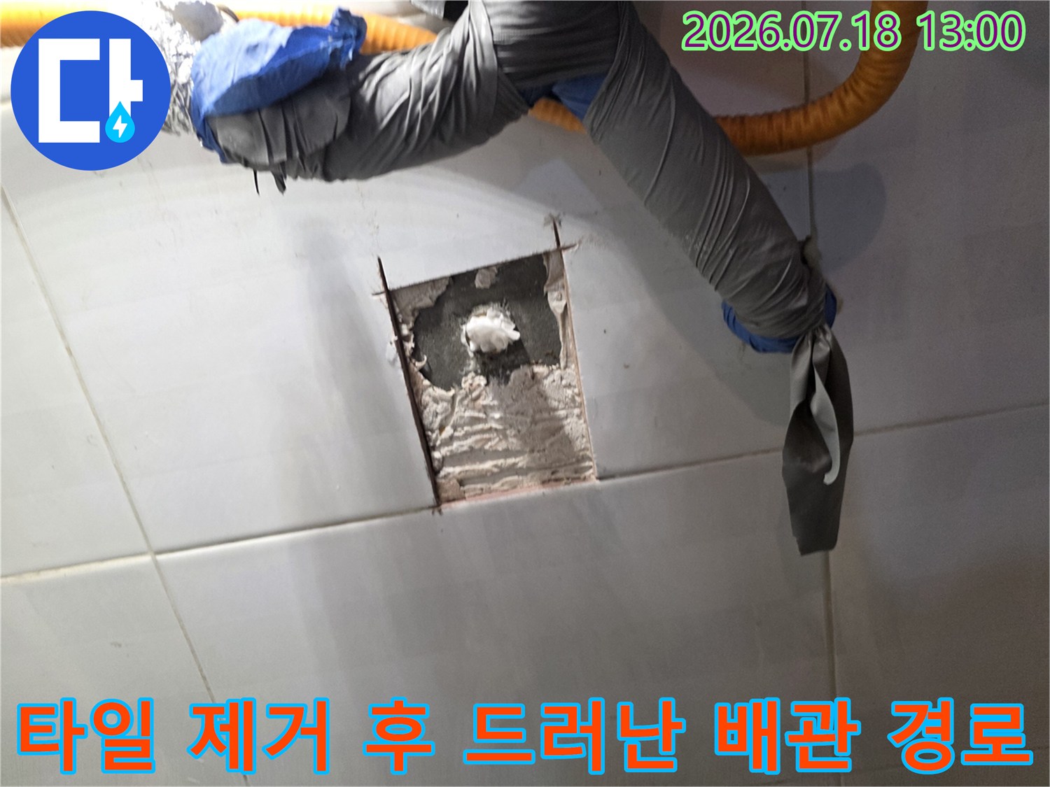 흰색 타일 벽면의 일부를 사각형으로 파내어 내부 시멘트 층이 보이고 상단에 회색과 파란색 보온재 배관이 있는 모습