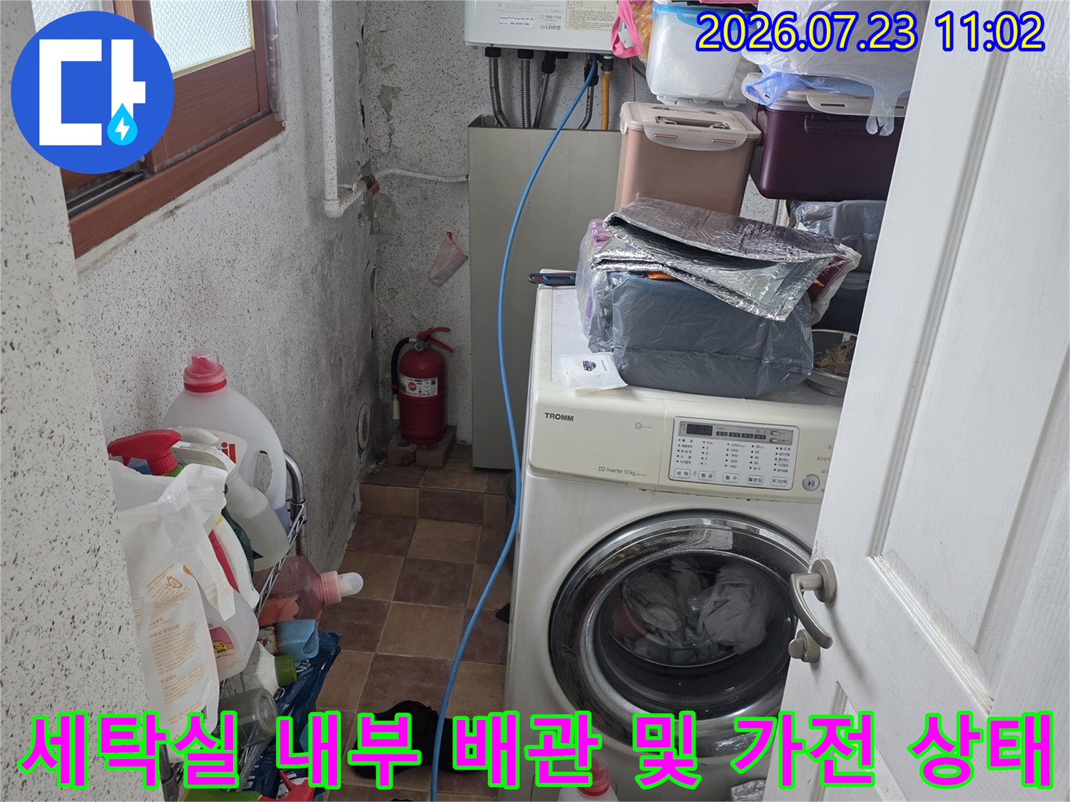 세탁기와 보일러가 설치된 다용도실-바닥에 놓인 세제들과 파란색 급수 호스 모습