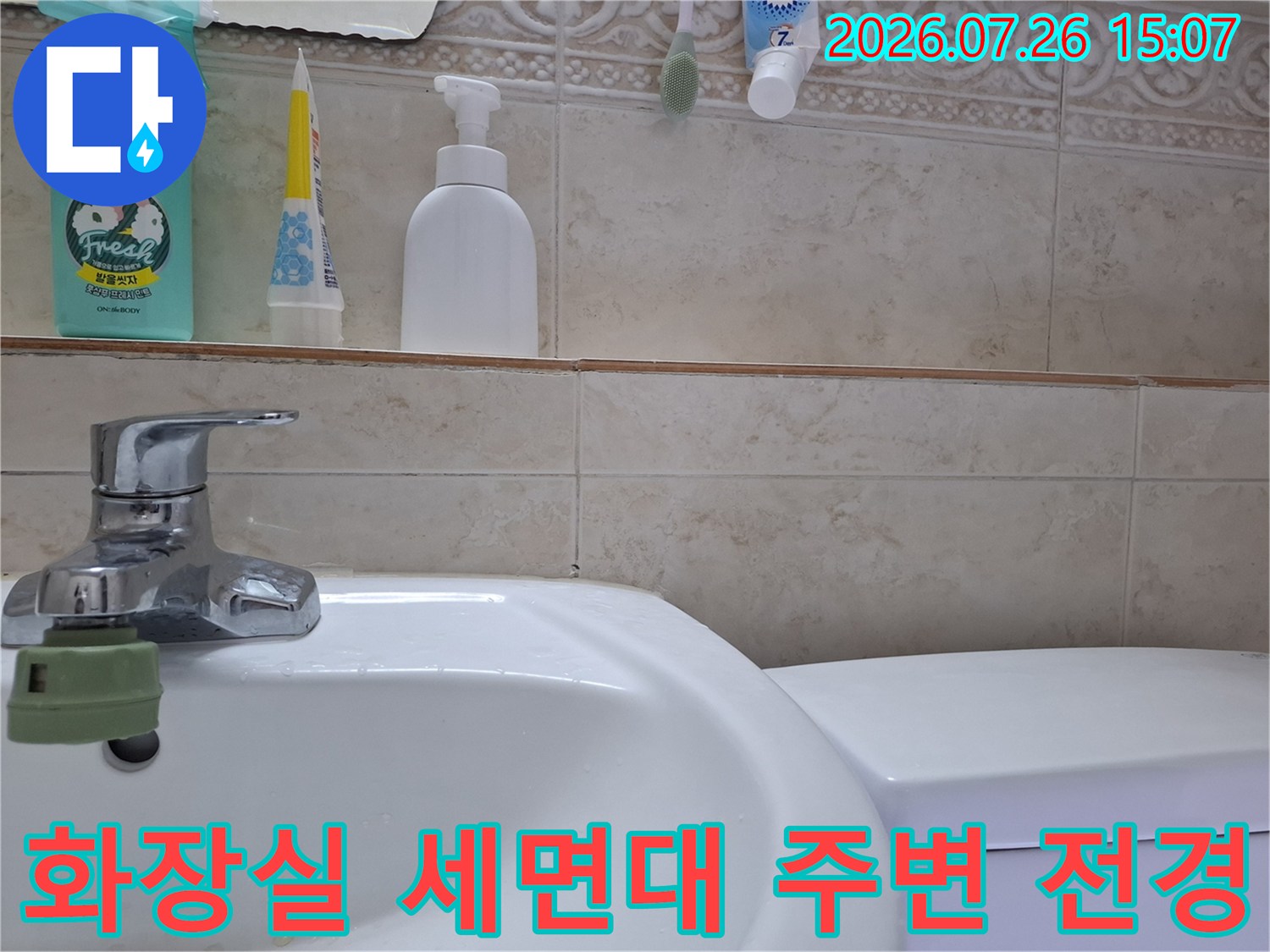 베이지색 타일이 시공된 욕실의 세면대와 수전-상단 선반에 놓인 여러 가지 위생 용품들
