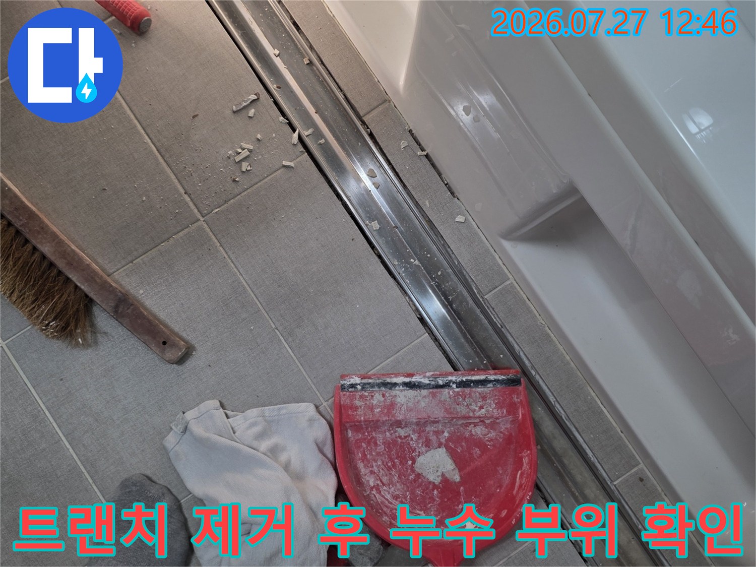 화장실 타일 바닥에 설치된 긴 배수구가 분리되어 있고 작업용 걸레와 도구들이 흩어져 있는 현장 사진
