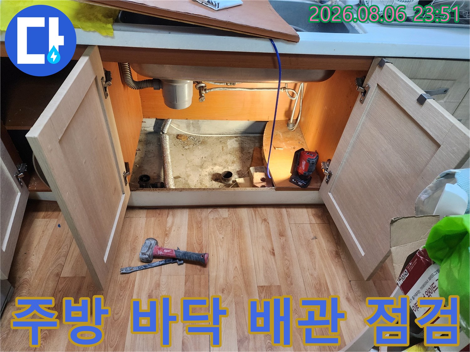 싱크대 하부장 내부의 노출된 배관과 작업 도구를 촬영한 사진입니다.