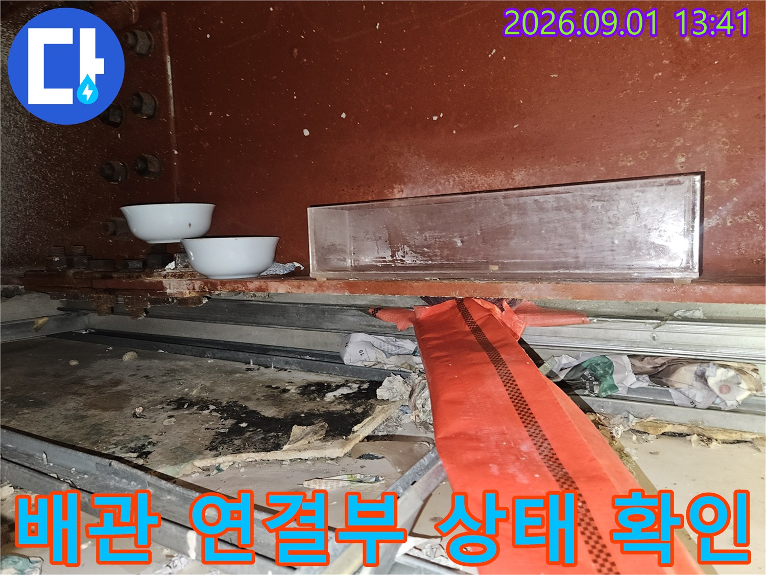 붉은색 벽면 아래 배관 연결 부위와 흰색 용기를 배치하여 누수 지점을 점검하는 모습입니다.
